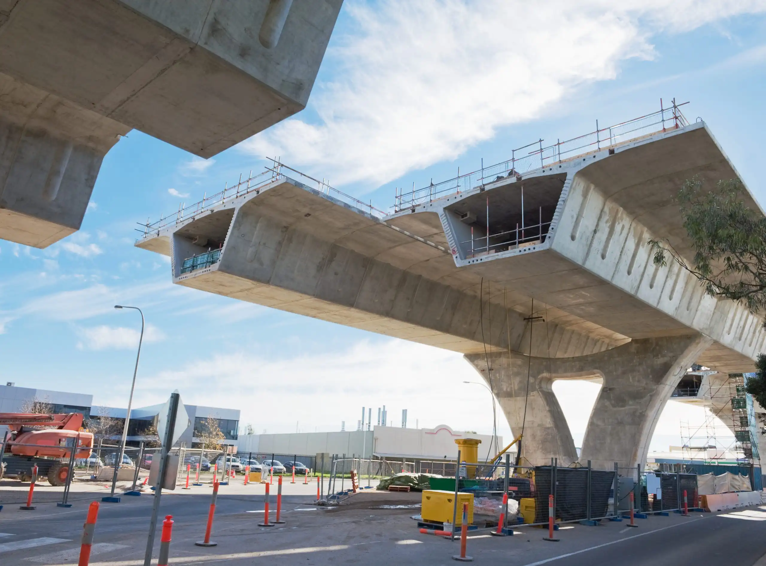 Concrete bridge structure under construction, featuring support beams, scaffolding, and traffic barriers against a clear blue sky.